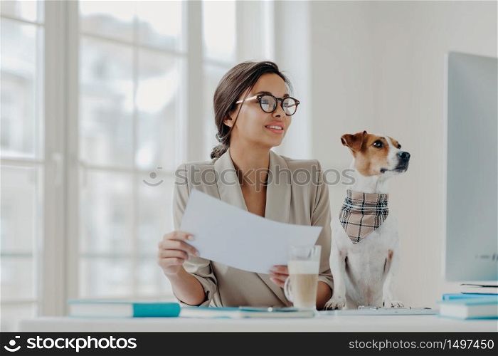 Photo of happy businesswoman works from home on self isolation, holds papers, checks information on computer, wears glasses, favorite pet poses near. Full concentration on work. Freelance worker