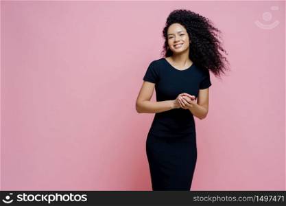 Photo of happy cheerful dark skinned curly woman dresses for casual event, smiles positively, enjoys pleasant talk with man, keeps hands together, stands indoor, free space aside for your text