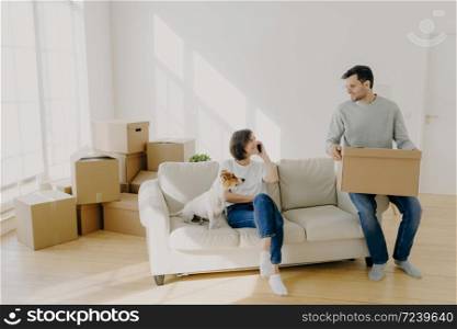 Photo of happy couple move in new home, pose on sofa with pet and boxes, have relocation, unpack boxes, female calls delivery service, carry belongings in carton containers. Welcome to new apartment