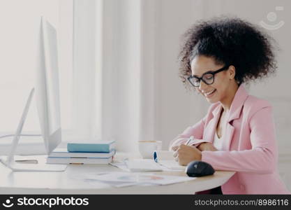 Photo of happy curly businesswoman dressed in elegant rosy suit, writes down something in notepad with cheerful expression, uses computer for searching information online, studies or works indoor