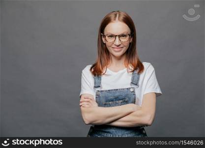 Photo of happy dark haired young woman keeps arms folded, smiles positively, wears eyewear, t shirt and denim overalls, isolated over grey background with free space for your information or text