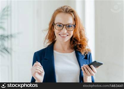 Photo of happy female leader with ginger hair, wears spectacles, updates app on modern cell phone, wears elegant clothes, poses in modern office, reads incoming message from business partner