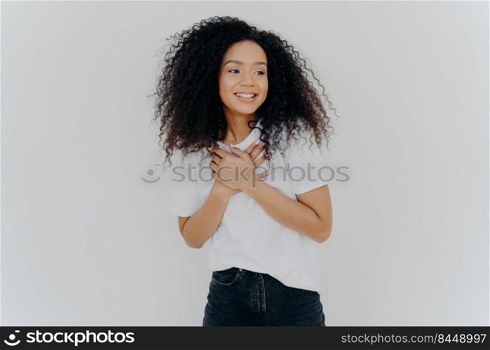 Photo of happy girl keeps both hands on chest, looks aside with p≤asant smi≤, notices something awesome, poses against white background, wears casual t shirt and jeans. Positive emotions concept