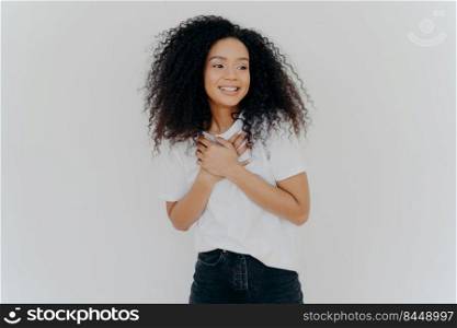 Photo of happy girl keeps both hands on chest, looks aside with pleasant smile, notices something awesome, poses against white background, wears casual t shirt and jeans. Positive emotions concept