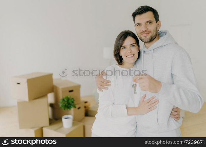 Photo of happy husband and wife buy real estate, cuddle and hold keys, stand in living room with boxes in new home, dressed in casual domestic clothes, receive keys from realtor. Relocation, mortgage