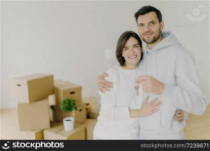 Photo of happy husband and wife buy real estate, cuddle and hold keys, stand in living room with boxes in new home, dressed in casual domestic clothes, receive keys from realtor. Relocation, mortgage