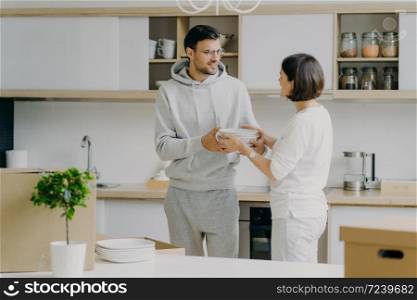 Photo of happy husband and wife unpack things after moving into new house, carry white plates, pose against kitchen interior, have fun, dressed in domestic clothes. People and moving concept