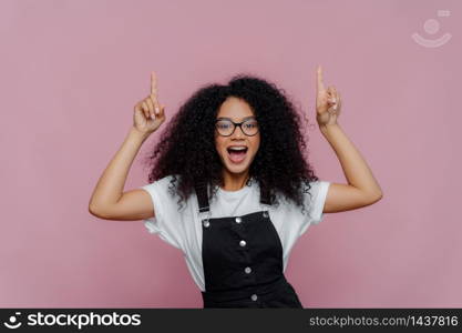 Photo of happy impressed Afro American woman points both index finger above, demonstrates awesome copy space, wears optical glasses and fashionable clothes, poses over violet background. Look above