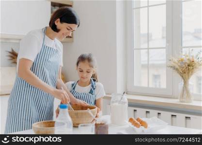 Photo of happy mum and child cook together at kitchen, wears aprons prepare something tasty, make food, whisk eggs with beater, bake house pastries at home. Milk, eggs, chocolate, flour on table