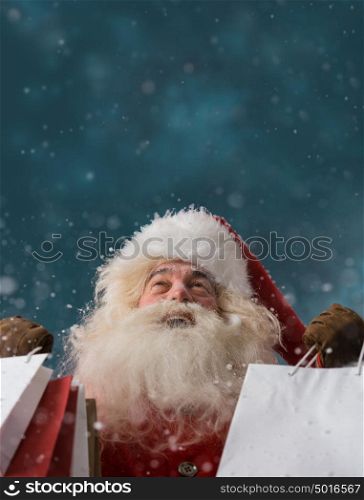 Photo of happy Santa Claus outdoors in snowfall holding shopping bags and looking upwards on big copyspace. Christmas sales and discount concept