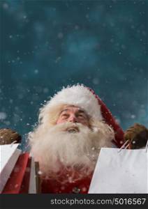 Photo of happy Santa Claus outdoors in snowfall holding shopping bags and looking upwards on big copyspace. Christmas sales and discount concept