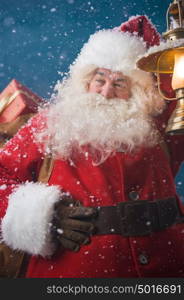 Photo of happy Santa Claus outdoors in snowfall lights the way with vintage lantern while carrying gifts to children