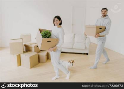 Photo of happy smiling brunette woman and her husband carry cardboard boxes with personal belongings, relocare to new apartment, enter empty room, start new life, little dog near, enjoy moving day