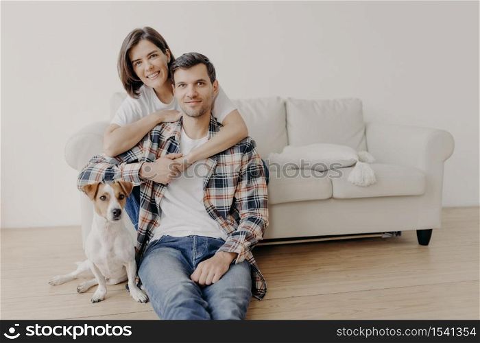 Photo of happy woman embraces her husband, poses on sofa, little dog sits on floor near owner, look all in camera with happy expressions, have romantic relationship. Family portrait. People and home