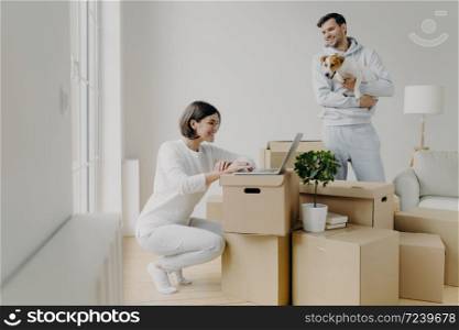Photo of happy woman works remotely on laptop computer, has happy smile, glad husband in casual clothes plays with pet, spend free time at their new home, surrounded with cardboard boxes in middle