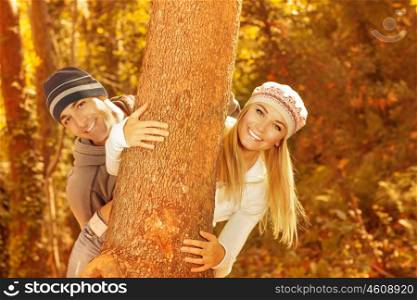 Photo of happy young family having fun in autumnal woods, closeup portrait of cute cheerful couple peeking from behind a tree outdoors, beautiful golden autumn season, love concept&#xA;