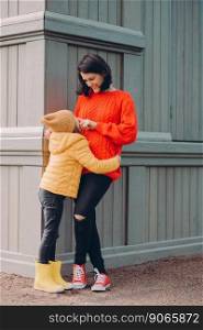 Photo of happy young mum touches nose of her curious pleased daughter, stand closely to each other at corner of wooden building. Lovely small child wears hat, jacket and boots, has fun with mother