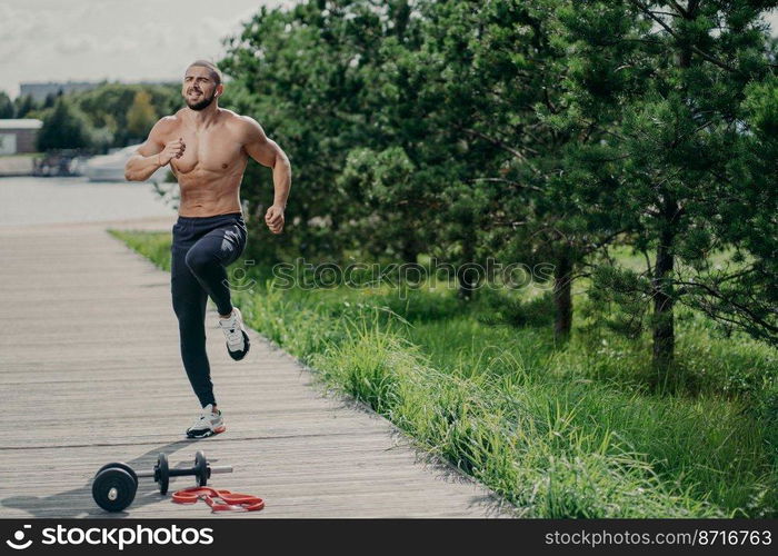 Photo of healthy motivated man with thick bristle, exercises outdoor, jumps and leads healthy lifestyle, poses near sport equipment, has strong muscular body. Fitness, sport and exercising concept