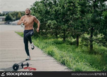 Photo of healthy motivated man with thick bristle, exercises outdoor, jumps and leads healthy lifestyle, poses near sport equipment, has strong muscular body. Fitness, sport and exercising concept