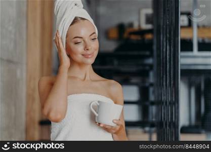 Photo of healthy thoughtful woman with healthy smooth skin drinks hot tea concentrated down, stands wrapped in white bath towel in cozy room, enjoys hygiene and beauty treatments, feels relaxed