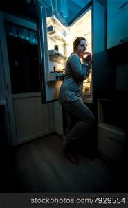 Photo of hungry woman eating at night near refrigerator