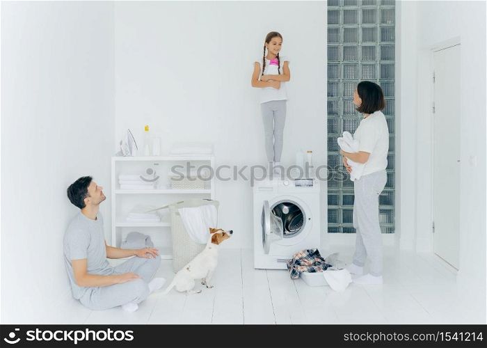Photo of husband and wife, their dog and daughter do housework in laundry room, little girl stands with detergent on top of washing machine, busy washing clothes and linen, have spring cleaning