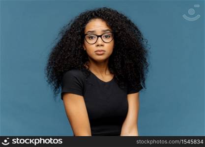 Photo of indignant puzzled Afro American woman looks surprisingly at camera, wears casual black t shirt and spectacles, poses against blue background. Human facial expressions and feelings concept