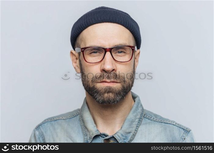 Photo of intelligent confident stylish man with dark thick beard and mustache, looks seriously into camera, poses against grey studio background, wears spectacles and hat. Facial expressions concept