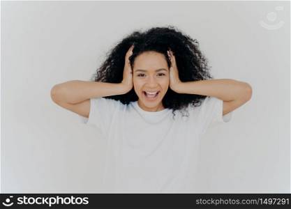 Photo of joyful dark skinned woman keeps hands on head, curly hair, laughs from joy, excited from positive emotions, dressed in white t shirt, hears funny joke, laughs sincerely. People, emotions
