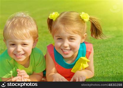 Photo of little brother and sister lying down on green grass field in sunny day, two adorable child playing on backyard, cute friends laughing outdoors in spring, preschooler kid, happy family concept