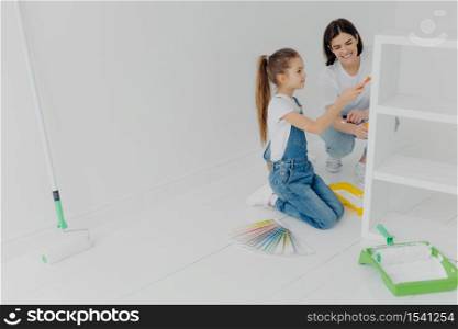 Photo of little girl and her mum paint furniture for new apartment, pose in empty room, use color samples, use painting tools, enjoy spending time together. House renovation and design concept
