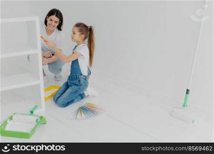 Photo of little girl and her mum paint furniture for new apartment, pose in empty room, use color samples, use painting tools, enjoy spending time together. House renovation and design concept