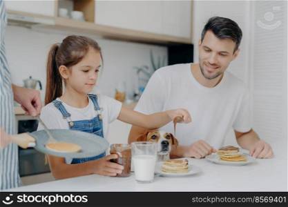 Photo of little girl in denim dungarees adds chocolate to pancakes, has breakfast together with dad and dog, likes how mother cooks. Family at kitchen have breakfast during weekend. Happy moment