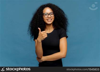 Photo of lovely dark skinned female makes like gesture, keeps thumb up, approves something good, has pleasant smile, wears spectacles and casual black t shirt, isolated over blue background.