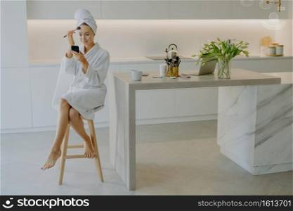 Photo of lovely female model applies foundation or powder on face with cosmetic brush looks in mirror poses at chair against kitchen interior wears white soft bathrobe and wrapped towel on head
