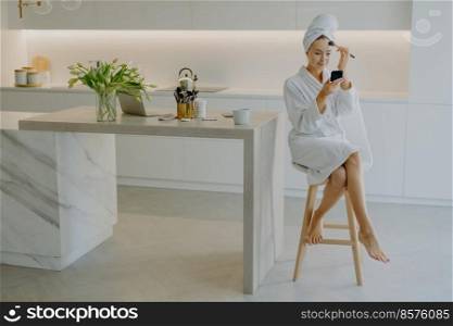 Photo of lovely female model applies foundation or powder on face with cosmetic brush looks in mirror poses at chair against kitchen interior wears white soft bathrobe and wrapped towel on head