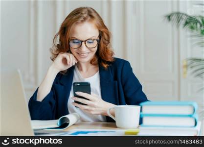Photo of lovely female sits with smartphone device, types feedback, works in office on up to date laptop, focused into screen of gadget, sits at work place with books, notepad and hot beverage