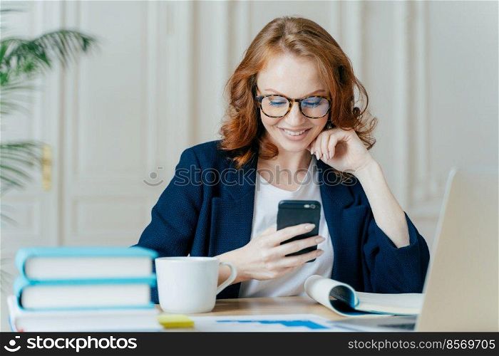 Photo of lovely female sits with smartphone device, types feedback, works in office on up to date laptop, focused into screen of gadget, sits at work place with books, notepad and hot beverage