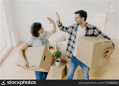 Photo of lovely husband and wife express their agreement, carry cardboard boxes with personal belongings, move in new apartment, pose in empty messy room, decide about interior of living room