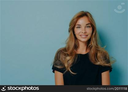 Photo of lovely long haired young woman looks directly at camera with tender expression dressed in casual black t shirt isolated over blue background blank copy space for your advertisement.. Long haired woman looks directly at camera with tender expression dressed in casual black t shirt