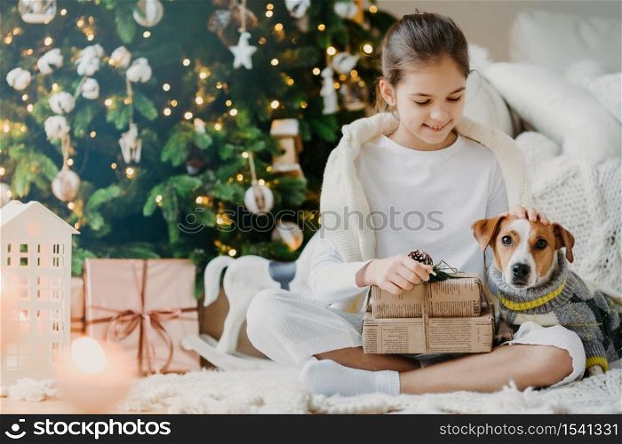 Photo of lovely small child sits crossed legs on floor, pets pedigree dog received Christmas presents from parents celebrate New Year together. Kid enjoys holiday with jack russell terrier opens gifts