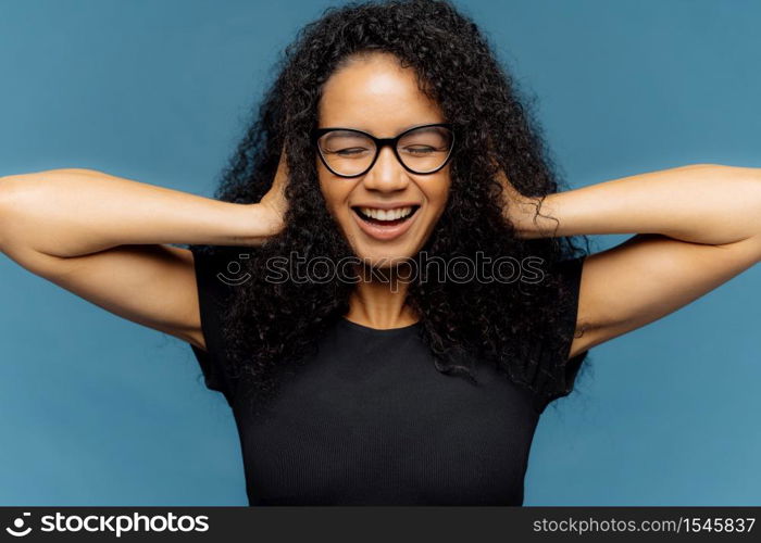 Photo of lovely smiling Afro female with crsip hair, covers ears, being in good mood, wears casual black t shirt, amused by loud music, isolated over blue background. People and positive emotions
