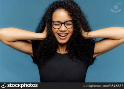 Photo of lovely smiling Afro female with crsip hair, covers ears, being in good mood, wears casual black t shirt, amused by loud music, isolated over blue background. People and positive emotions