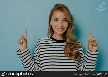 Photo of lovely smiling young woman points fingers up shows promo logo overhead store advertisement wears casual striped jumper isolated over blue background makes her choice. Promotion concept. Photo of lovely smiling young woman points fingers up shows promo logo overhead