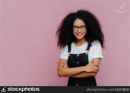 Photo of lovely teenage girl with Afro haircut, keeps arms folded, wears glasses, white t shirt and black overalls, looks gladfully at camera, stands against purple background with copy space for text