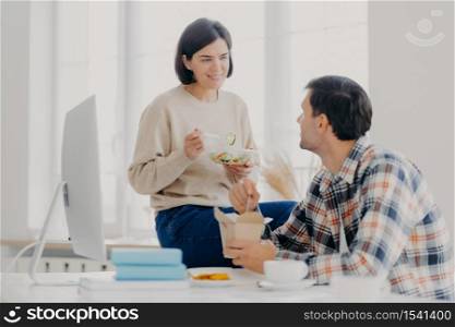 Photo of lovely woman and man discuss working issues together while have lunch, eat fast food and fresh vegetable salad, pose at desktop with computer monitor and scientific literature. Teamwork