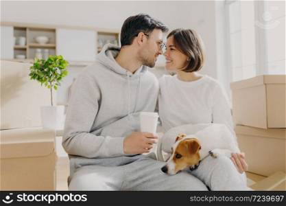 Photo of lovely woman and man going to kiss, husband holds takeaway coffee, sit indoor near cardboard boxes, unpack personal belongings on relocating day, live with pet, buy new expensive flat