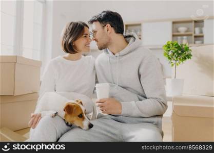 Photo of lovely woman and man going to kiss, husband holds takeaway coffee, sit indoor near cardboard boxes, unpack personal belongings on relocating day, live with pet, buy new expensive flat