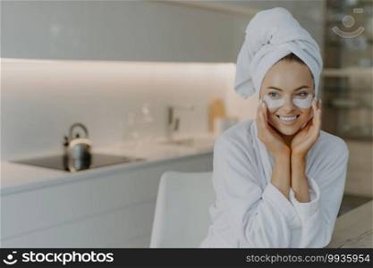 Photo of lovely woman with healthy skin applies recovery pads under eyes enjoys skin care and effective beauty treatment dressed in soft bathrobe wears wrapped towel on head poses over home interior