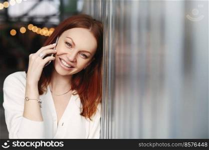 Photo of lovely woman with reddish hair, positive smile, has telephone talk with customer, calls to friend, dressed in white shirt, looks at camera with satisfied expression, enjoys communication